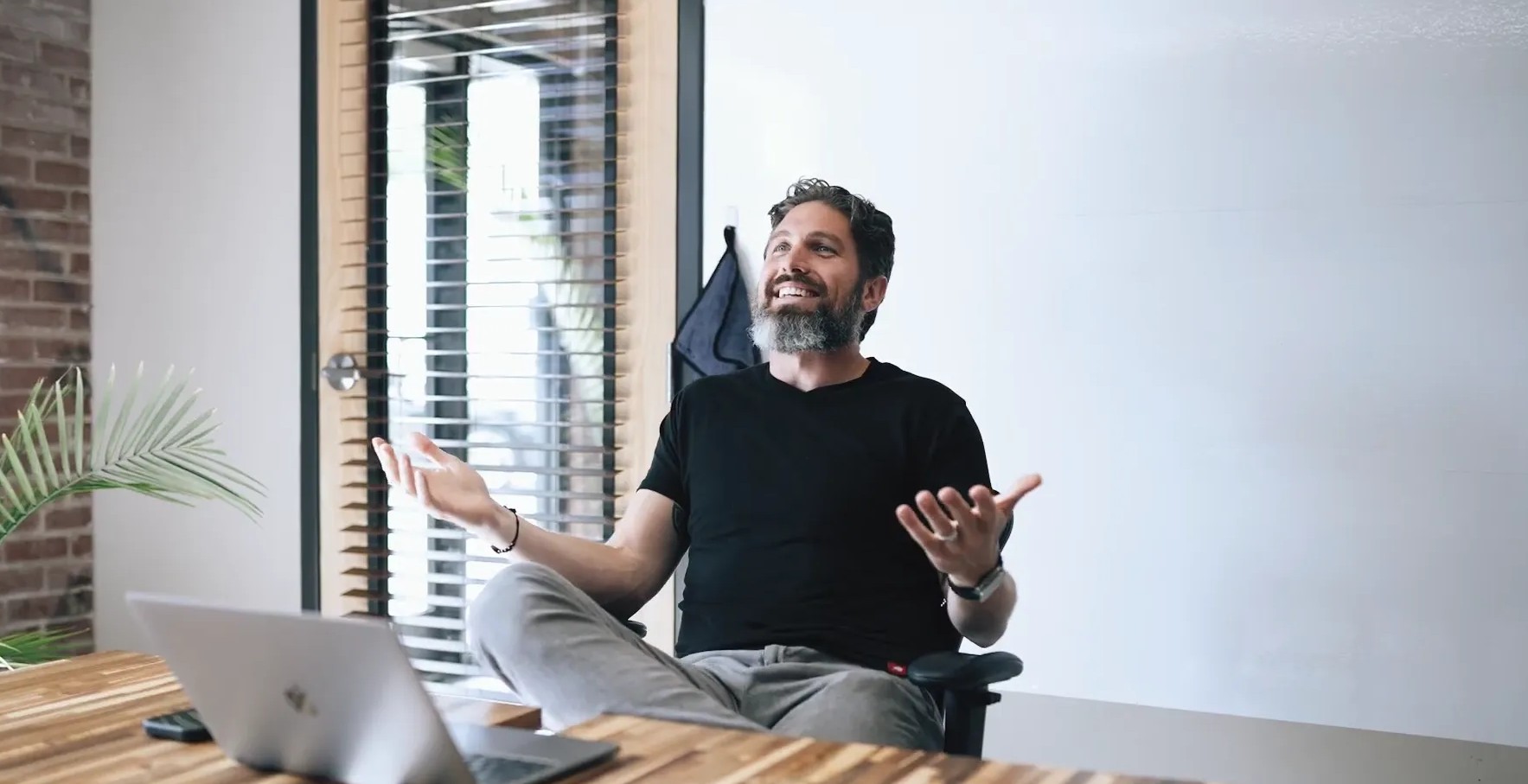 A man sits at a table gesturing while looking at a laptop.