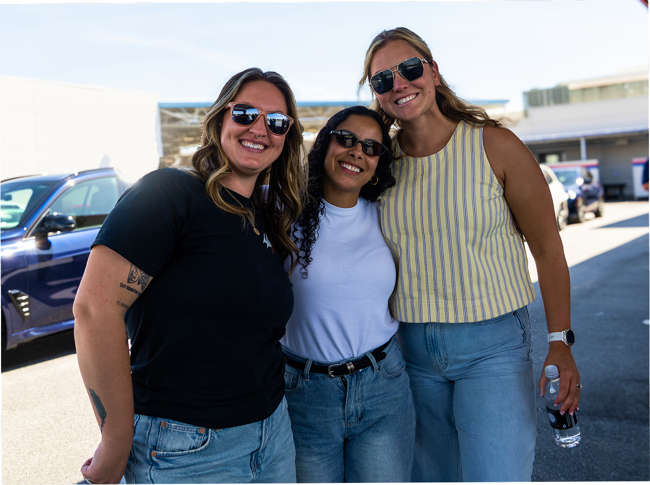 Three women stand outdoors smiling in front of a building.