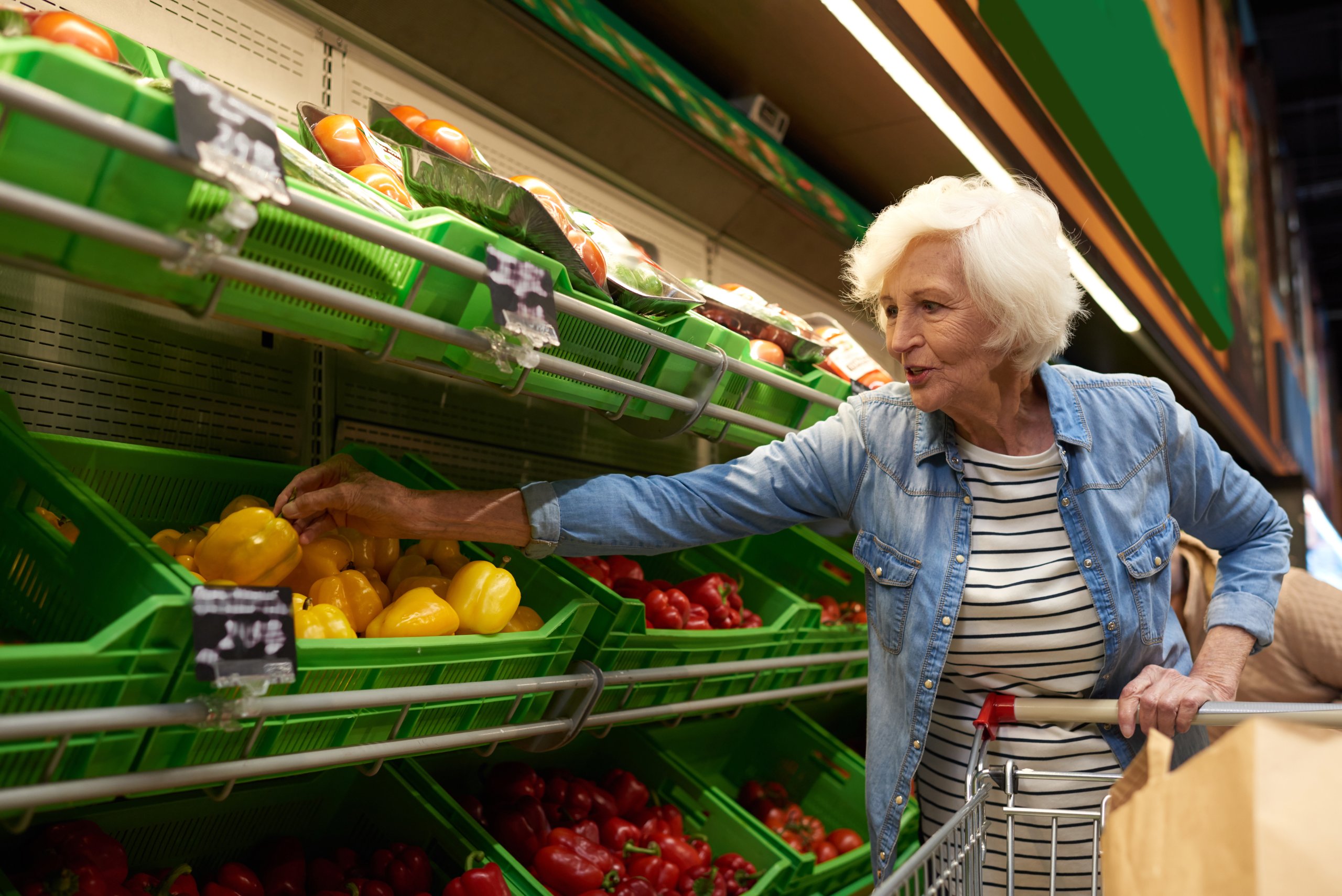 An elderly woman selects yellow and red vegetables in a grocery store.