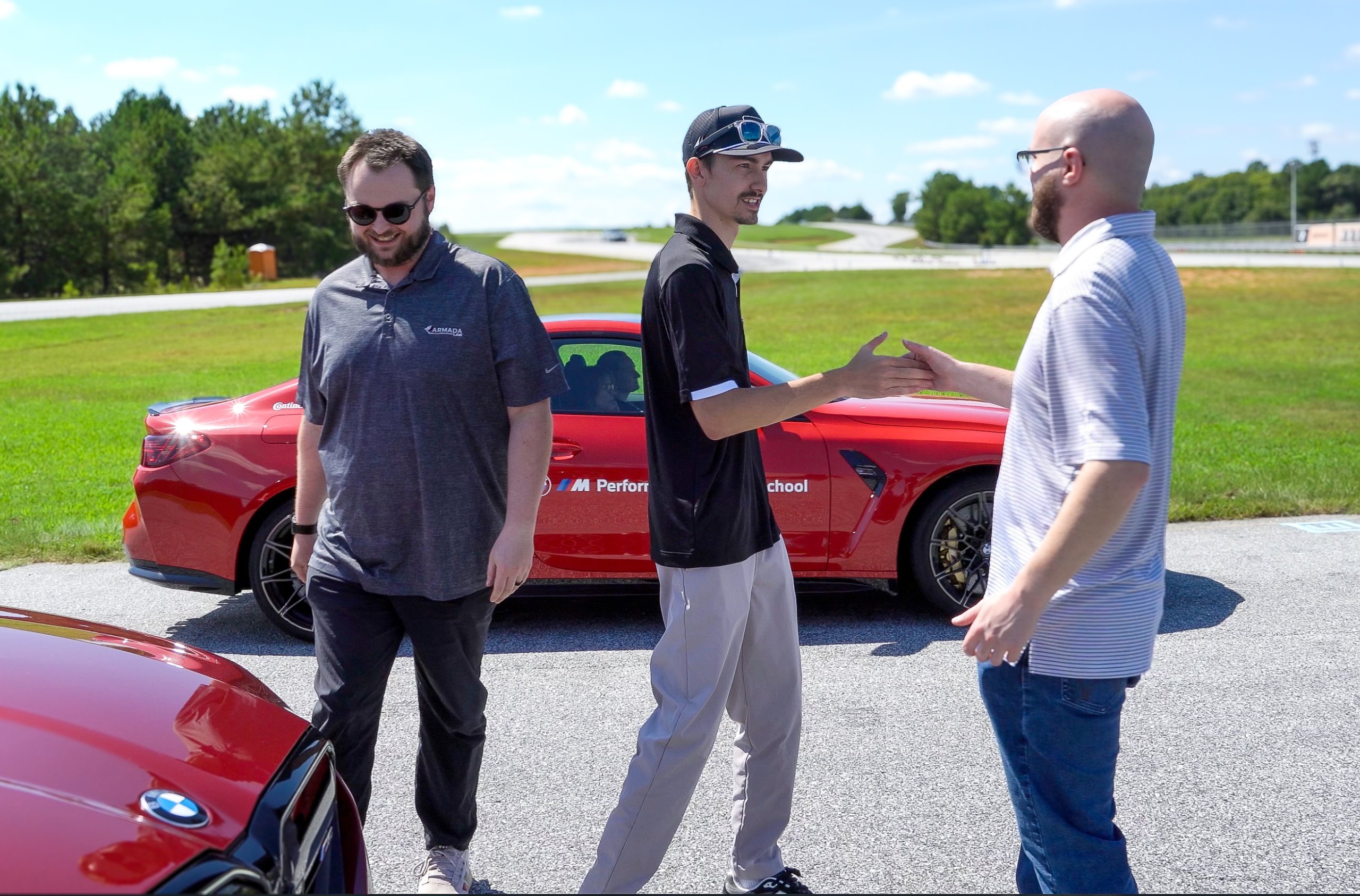 Three men stand near a red car outdoors.