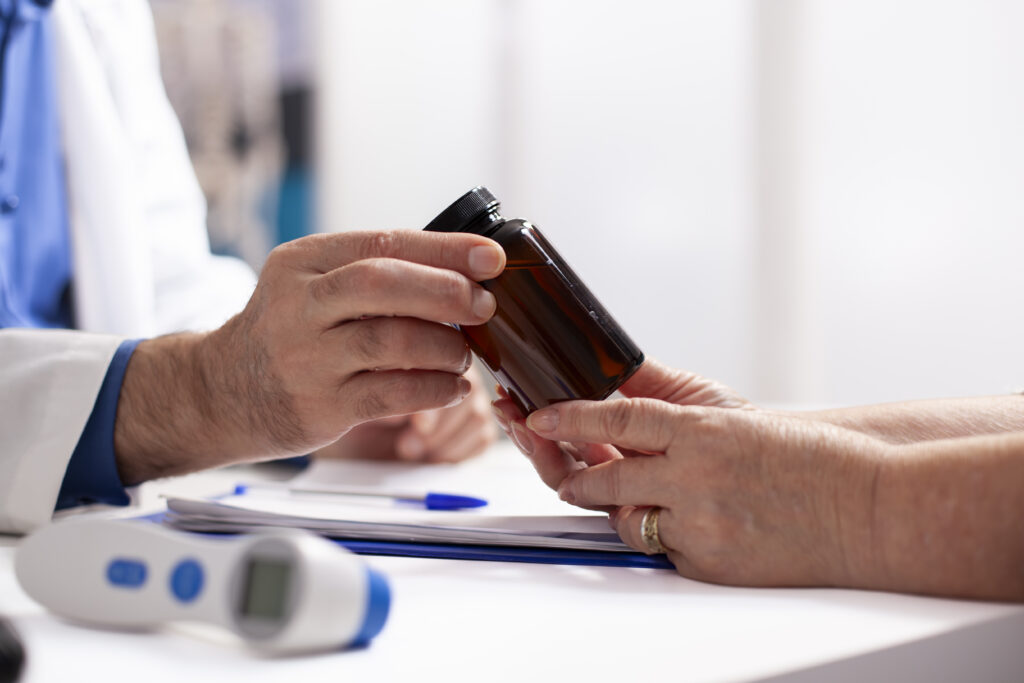 Closeup of person in lab coat giving medicine to pensioner patient after medical exams. Selective focus of caucasian people holding pill bottle on table with thermometer and clipboard nearby.